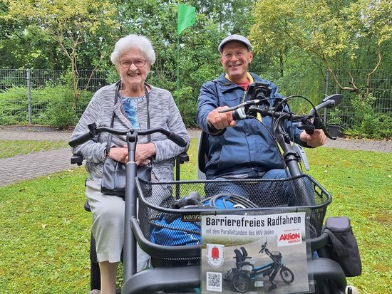 Zwei ältere Menschen sitzen lächelnd auf einem blauen Tandem-Fahrzeug im Park. Der Mann steuert, die Frau sitzt daneben. An der Front ist ein Schild mit der Aufschrift „Barrierefreies Radfahren“ angebracht.